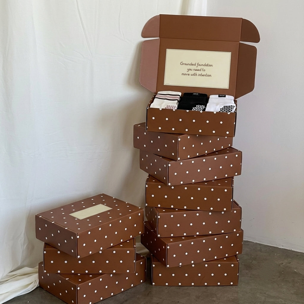 A stack of brown gift boxes with white polka dots, with one open box showing folded grippy socks inside. Another closed box sits on the floor beside it against a white draped backdrop.